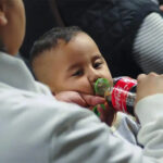 A mother standing next to a seated toddler boy in some kind of theater or auditorium. The mother is feeding the boy Coca-Cola from a bottle and holding his pacifier in her other hand.