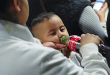 A mother standing next to a seated toddler boy in some kind of theater or auditorium. The mother is feeding the boy Coca-Cola from a bottle and holding his pacifier in her other hand.