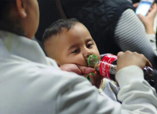 A mother standing next to a seated toddler boy in some kind of theater or auditorium. The mother is feeding the boy Coca-Cola from a bottle and holding his pacifier in her other hand.