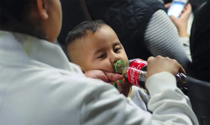 A mother standing next to a seated toddler boy in some kind of theater or auditorium. The mother is feeding the boy Coca-Cola from a bottle and holding his pacifier in her other hand.