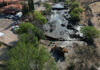 Heavy machinery sits in a river bed with a partly demolished dam, next to a large hacienda-style house