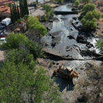 Heavy machinery sits in a river bed with a partly demolished dam, next to a large hacienda-style house