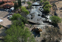 Heavy machinery sits in a river bed with a partly demolished dam, next to a large hacienda-style house