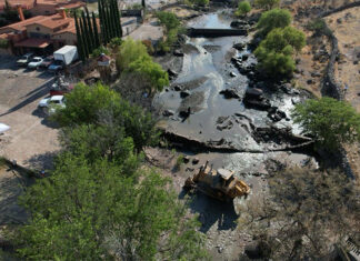 Heavy machinery sits in a river bed with a partly demolished dam, next to a large hacienda-style house