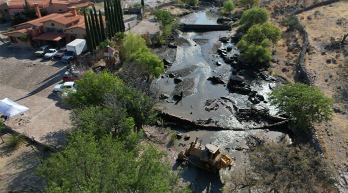 Heavy machinery sits in a river bed with a partly demolished dam, next to a large hacienda-style house