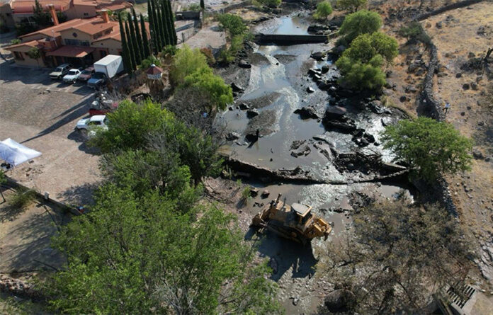 Heavy machinery sits in a river bed with a partly demolished dam, next to a large hacienda-style house