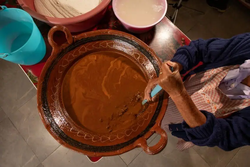 An aerial view of an elderly Mexican woman in a traditional apron and a blue cardigan stirring a large, traditional clay bowl of mole poblano.