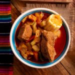 A bowl of mole de olla displayed close up in the center of a table in a food photography style. On the table are also a potholder made of natural fibers and a traditional woven Mexican textile featuring multicolor thin striping.