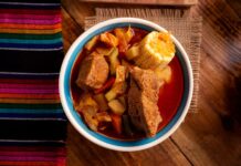 A bowl of mole de olla displayed close up in the center of a table in a food photography style. On the table are also a potholder made of natural fibers and a traditional woven Mexican textile featuring multicolor thin striping.