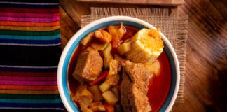A bowl of mole de olla displayed close up in the center of a table in a food photography style. On the table are also a potholder made of natural fibers and a traditional woven Mexican textile featuring multicolor thin striping.