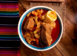 A bowl of mole de olla displayed close up in the center of a table in a food photography style. On the table are also a potholder made of natural fibers and a traditional woven Mexican textile featuring multicolor thin striping.