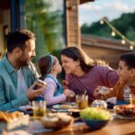 Stock image of a young mother and father at a meal outdoors at a wooden table filled with serving plates of food. They are interacting with their young children. The mother is squinting playfully at her daughter in her partner's arms. The daughter holds her face up close to her mother's while the young son looks on from her other side.
