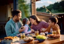 Stock image of a young mother and father at a meal outdoors at a wooden table filled with serving plates of food. They are interacting with their young children. The mother is squinting playfully at her daughter in her partner's arms. The daughter holds her face up close to her mother's while the young son looks on from her other side.