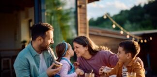Stock image of a young mother and father at a meal outdoors at a wooden table filled with serving plates of food. They are interacting with their young children. The mother is squinting playfully at her daughter in her partner's arms. The daughter holds her face up close to her mother's while the young son looks on from her other side.