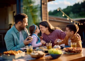 Stock image of a young mother and father at a meal outdoors at a wooden table filled with serving plates of food. They are interacting with their young children. The mother is squinting playfully at her daughter in her partner's arms. The daughter holds her face up close to her mother's while the young son looks on from her other side.