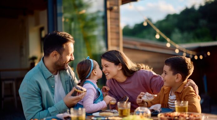Stock image of a young mother and father at a meal outdoors at a wooden table filled with serving plates of food. They are interacting with their young children. The mother is squinting playfully at her daughter in her partner's arms. The daughter holds her face up close to her mother's while the young son looks on from her other side.