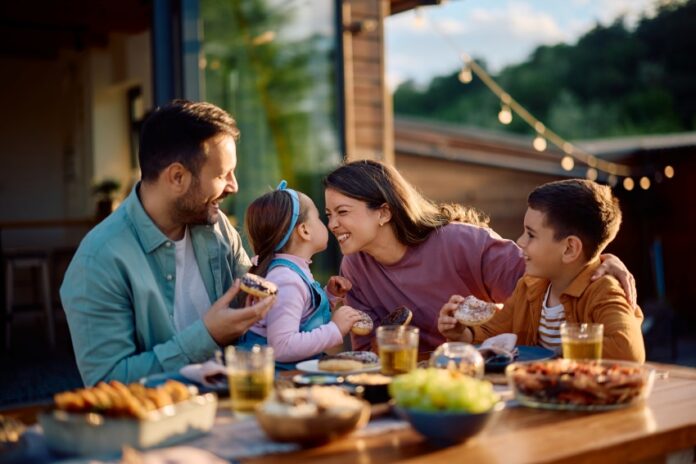 Stock image of a young mother and father at a meal outdoors at a wooden table filled with serving plates of food. They are interacting with their young children. The mother is squinting playfully at her daughter in her partner's arms. The daughter holds her face up close to her mother's while the young son looks on from her other side.