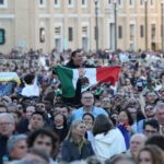A crowd in St. Peter's Square watching the first public address of Robert Prevost as Pope Leo XIV. Above their shoulders, an exuberant Mexican man holds a Mexican flag in front of his chest as he watches.