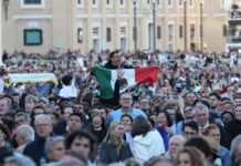 A crowd in St. Peter's Square watching the first public address of Robert Prevost as Pope Leo XIV. Above their shoulders, an exuberant Mexican man holds a Mexican flag in front of his chest as he watches.
