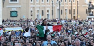 A crowd in St. Peter's Square watching the first public address of Robert Prevost as Pope Leo XIV. Above their shoulders, an exuberant Mexican man holds a Mexican flag in front of his chest as he watches.