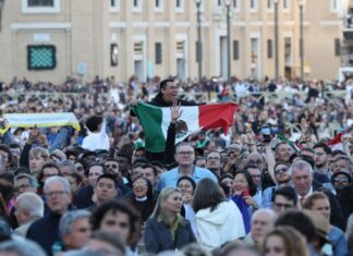 A crowd in St. Peter's Square watching the first public address of Robert Prevost as Pope Leo XIV. Above their shoulders, an exuberant Mexican man holds a Mexican flag in front of his chest as he watches.