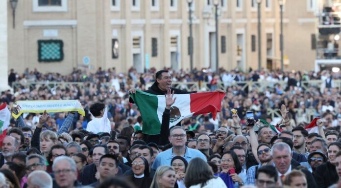 A crowd in St. Peter's Square watching the first public address of Robert Prevost as Pope Leo XIV. Above their shoulders, an exuberant Mexican man holds a Mexican flag in front of his chest as he watches.