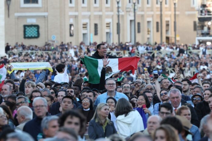 A crowd in St. Peter's Square watching the first public address of Robert Prevost as Pope Leo XIV. Above their shoulders, an exuberant Mexican man holds a Mexican flag in front of his chest as he watches.