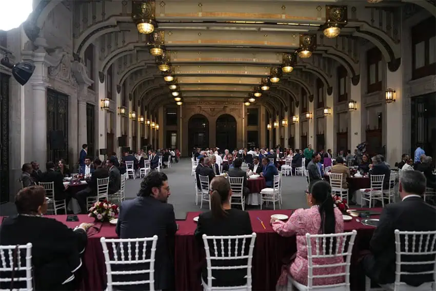 Sheinbaum and officials sit at tables on a dias in a room filled with guests seated at tables
