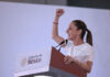 Mexican President Claudia Sheinbaum raising her arm while standing at a podium with the sigil of the Mexican government. She is looking out to an audience off camera. She is delivering Baja California Sur news.