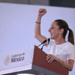 Mexican President Claudia Sheinbaum raising her arm while standing at a podium with the sigil of the Mexican government. She is looking out to an audience off camera. She is delivering Baja California Sur news.