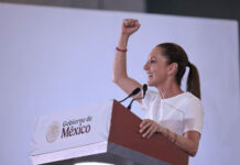 Mexican President Claudia Sheinbaum raising her arm while standing at a podium with the sigil of the Mexican government. She is looking out to an audience off camera. She is delivering Baja California Sur news.