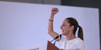 Mexican President Claudia Sheinbaum raising her arm while standing at a podium with the sigil of the Mexican government. She is looking out to an audience off camera. She is delivering Baja California Sur news.