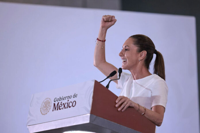 Mexican President Claudia Sheinbaum raising her arm while standing at a podium with the sigil of the Mexican government. She is looking out to an audience off camera. She is delivering Baja California Sur news.