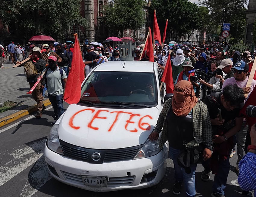 Masked teachers union protesters march down a street surrounding a car with the letters CETEG spray painted on it