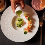 A fine dining presentation of a dish involving pieces of sushi and fresh fruit on a white plate on a placemat next to a cocktail and tortillas on an artisan stone bowl.