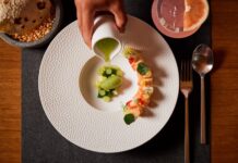 A fine dining presentation of a dish involving pieces of sushi and fresh fruit on a white plate on a placemat next to a cocktail and tortillas on an artisan stone bowl.
