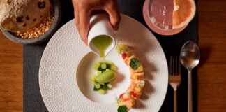 A fine dining presentation of a dish involving pieces of sushi and fresh fruit on a white plate on a placemat next to a cocktail and tortillas on an artisan stone bowl.