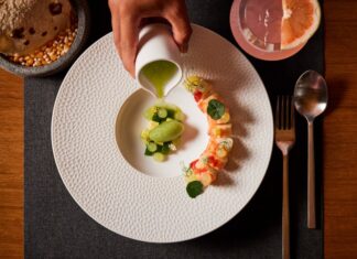 A fine dining presentation of a dish involving pieces of sushi and fresh fruit on a white plate on a placemat next to a cocktail and tortillas on an artisan stone bowl.
