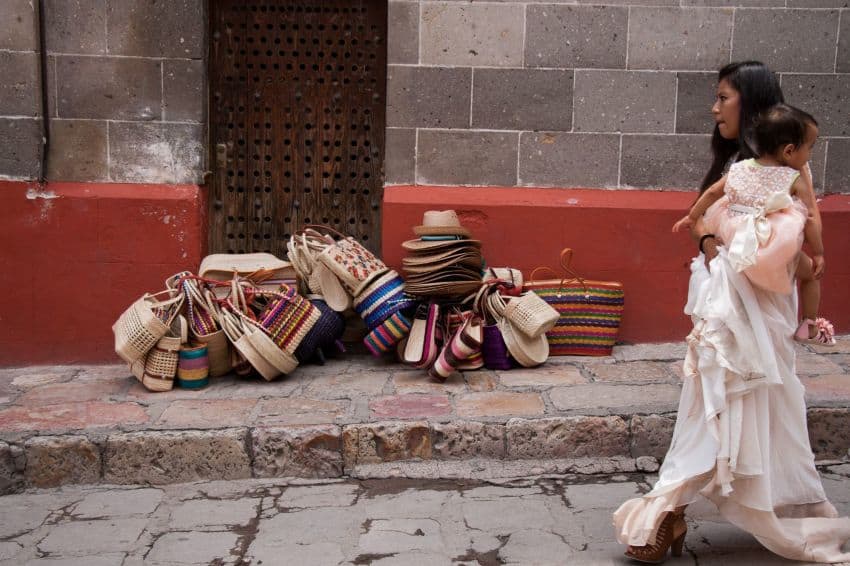 A young Mexican woman in a white dress and platform heeled shoes walks with her toddler daughter in a white dress in her arms down a stone paved street in San Miguel de Allende, Mexico.