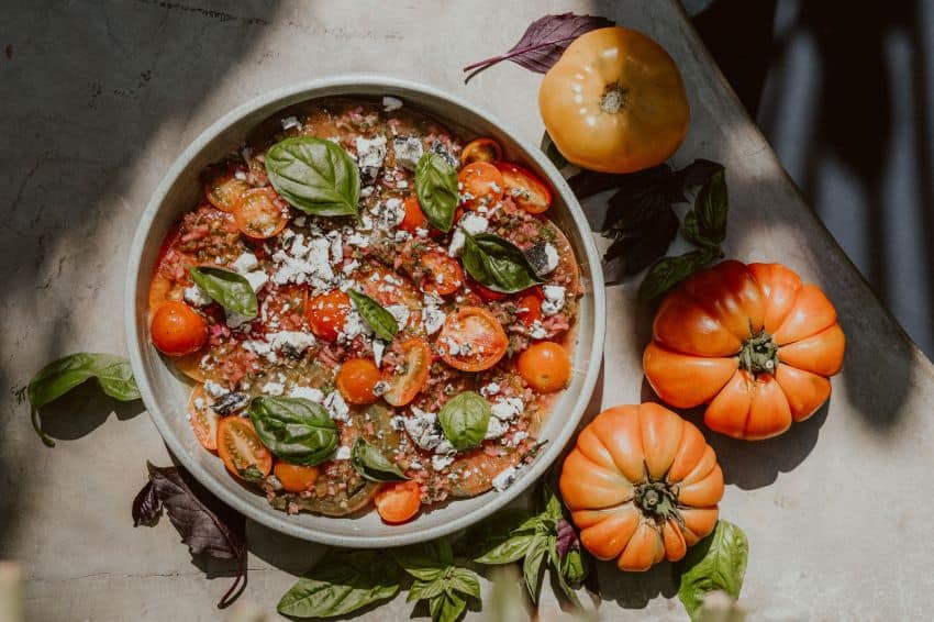 Roasted tomatoes artfully displayed on a serving plate with leaves of fresh basil, surrounded by fat heirloom tomatoes.