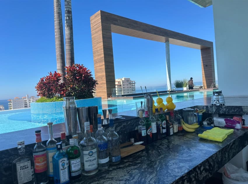 A bar with liquor bottles and blenders at a rooftop bar surrounded bya pool.