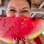 Woman posing in a Mexican palapa structure with a wedge of ripe watermelon in front of her face.