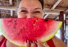 Woman posing in a Mexican palapa structure with a wedge of ripe watermelon in front of her face.