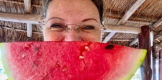 Woman posing in a Mexican palapa structure with a wedge of ripe watermelon in front of her face.