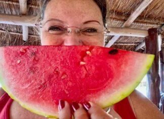 Woman posing in a Mexican palapa structure with a wedge of ripe watermelon in front of her face.