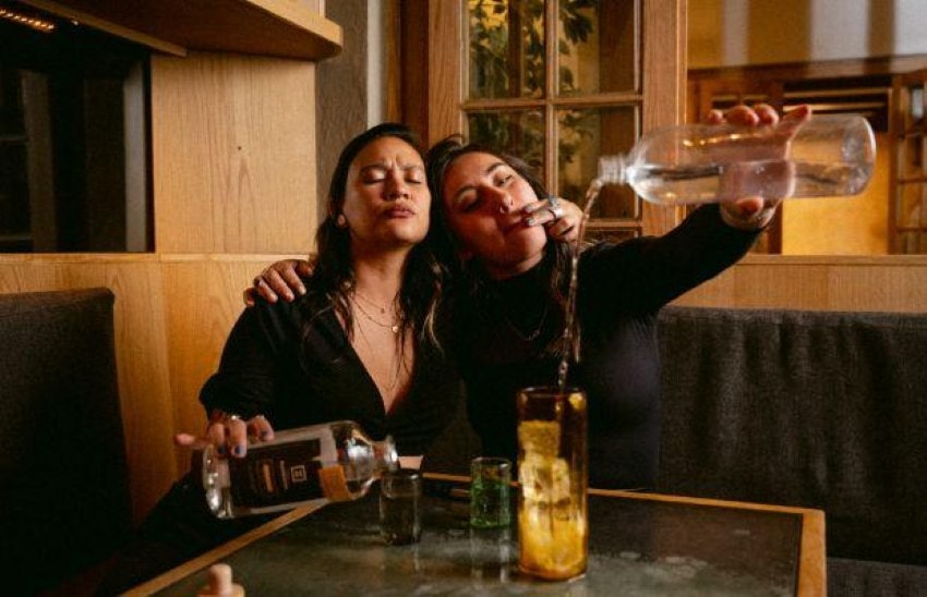 Two women in black blouses sitting at a cocktail bar table, sitting side-by-side, one with her arm around the other one's shoulder. Each holds a bottle of unspecified liquor in their hands that they are pouring into a glass.