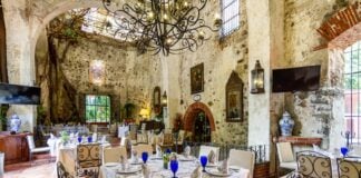 An elegant dining room with a few tables surrounded by original stone walls of the historic Mexican hacienda it occupies. There are wrought iron chandeliers hanging from the ceiling, a stone tile floor and traditional Mexican paintings and decor everywhere.