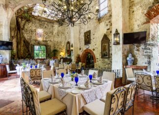 An elegant dining room with a few tables surrounded by original stone walls of the historic Mexican hacienda it occupies. There are wrought iron chandeliers hanging from the ceiling, a stone tile floor and traditional Mexican paintings and decor everywhere.