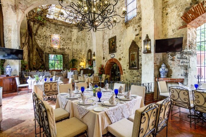 An elegant dining room with a few tables surrounded by original stone walls of the historic Mexican hacienda it occupies. There are wrought iron chandeliers hanging from the ceiling, a stone tile floor and traditional Mexican paintings and decor everywhere.