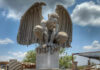 A large, light-colored cantera stone sculpture of a crouching gargoyle with outstretched wings, set against a blue sky with some clouds.